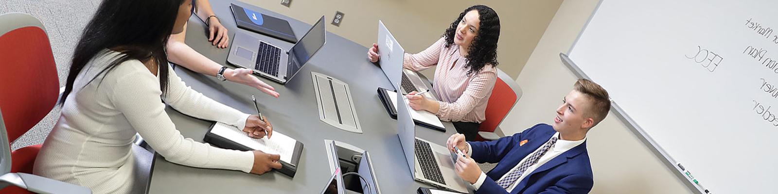 Students at conference room table with laptops, white board, and large display monitor